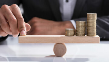 Close-up of A Businessperson's Hand Balancing Stacked Coins On Wooden Seesaw With Finger Over Desk